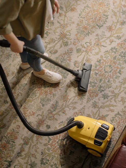 A person using a vacuum cleaner with a yellow body and black hose to deep clean a patterned area rug in a residential living room. The rug features beige, green, and orange floral designs on a cream background. The individual, wearing beige sneakers, blue jeans, and a beige jacket, is holding the vacuum's handle, moving it across the rug. The room's lighting highlights the clean, well-maintained surface, and the surrounding floor and furniture are visible. This image showcases surface cleaning and deep cleaning processes provided by Cleaners Surrey, illustrating professional carpet cleaning for domestic spaces.