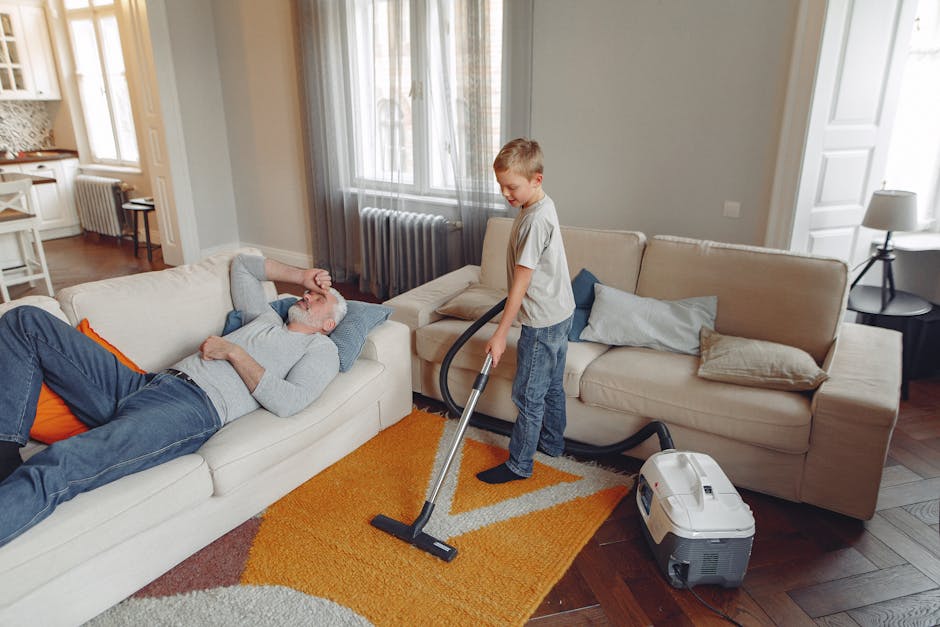 A young boy is vacuuming a beige fabric sofa in a living room with light grey walls, large windows with sheer curtains, and a wooden floor covered partially by an orange and beige patterned rug. An older man with grey hair and a beard is lying on the sofa, resting with his eyes closed. The boy is holding a modern vacuum cleaner with a hose attachment, and the vacuum unit is positioned on the floor nearby. The room is well-lit with natural light, and the setting appears clean and tidy, demonstrating a domestic cleaning process. This image illustrates the use of surface cleaning tools in a residential space, with Cleaners Surrey providing professional deep cleaning services to ensure hygienic and maintained home environments.
