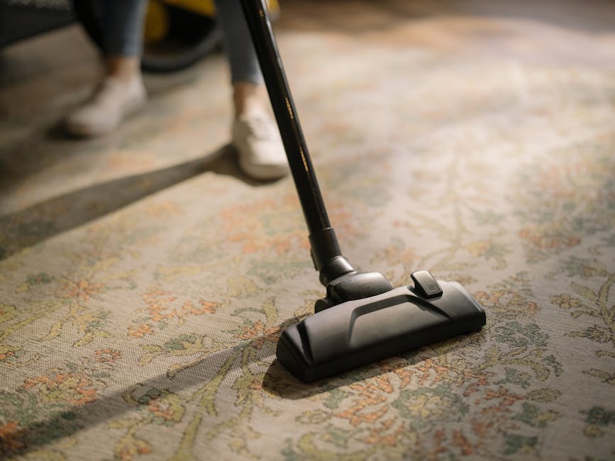A person using a vacuum cleaner with a yellow body and black hose to deep clean a patterned area rug in a residential living room. The rug features beige, green, and orange floral designs on a cream background. The individual, wearing beige sneakers, blue jeans, and a beige jacket, is holding the vacuum's handle, moving it across the rug. The room's lighting highlights the clean, well-maintained surface, and the surrounding floor and furniture are visible. This image showcases surface cleaning and deep cleaning processes provided by Cleaners Surrey, illustrating professional carpet cleaning for domestic spaces.