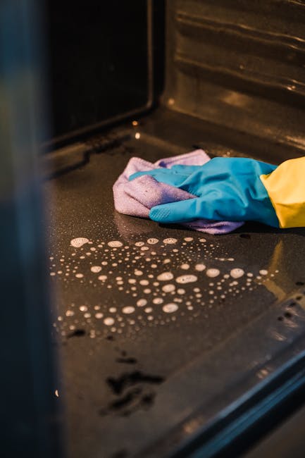 Close-up view inside an oven, showing a black ceramic or enamel surface with a worker wearing a yellow sleeve and blue gloves, using a pink and purple cleaning cloth to wipe away grease and residue. T