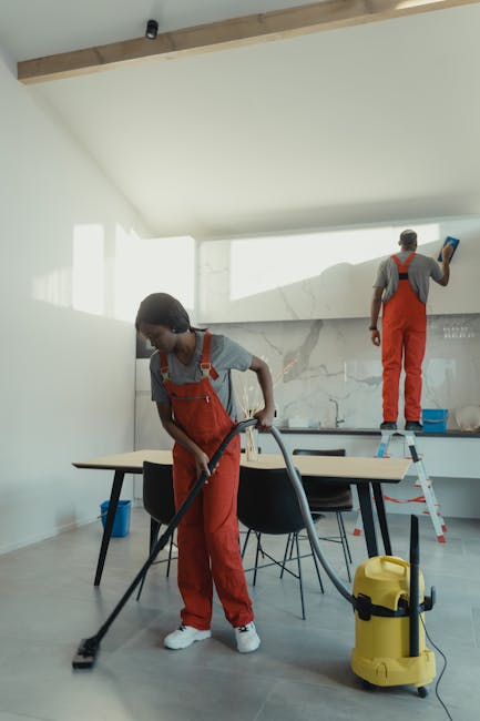 Two professional cleaners from Cleaners Surrey are engaged in a deep cleaning process within a modern kitchen area. One cleaner, positioned in the foreground, uses a vacuum cleaner on a tiled floor, which appears to be thoroughly dusted and free of debris, with the vacuum's cord extending toward the wall. The second cleaner stands on a step ladder at the back, wiping or cleaning the high kitchen cabinets or wall-mounted area with a cloth, suggesting a focus on surface sanitisation. The kitchen features a marble backsplash, a large wooden dining table with black chairs, and natural daylight flooding through large windows, enhancing the room's brightness and cleanliness. The surfaces, including the wooden table, marble, and tiled floor, look clean and well-maintained, reflecting professional deep cleaning practices. The overall scene emphasizes a comprehensive domestic cleaning service provided by Cleaners Surrey, tailored for end-of-tenancy or routine surface cleaning and sanitisation tasks.