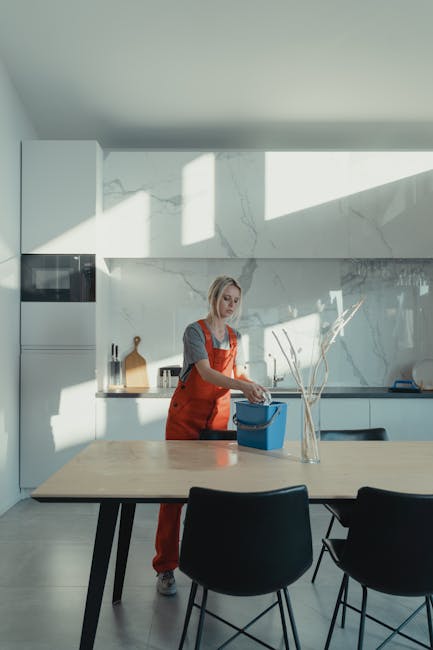 A female cleaner from Cleaners Surrey performing surface cleaning in a modern kitchen. She is dressed in an orange uniform and is using a spray bottle and cloth to wipe down a light-colored, polished wooden dining table. The kitchen features white marble countertops and backsplash, with stainless steel appliances integrated into the cabinetry. Natural light streams through a large window, casting shadows on the wall and creating a bright, calm atmosphere. The scene emphasizes domestic cleaning through thorough sanitisation and surface maintenance of the dining and kitchen surfaces, demonstrating hygienic cleaning processes in a residential setting. The space appears well-maintained and free of dust or stains, reflecting effective deep cleaning by professionals like those at Cleaners Surrey.