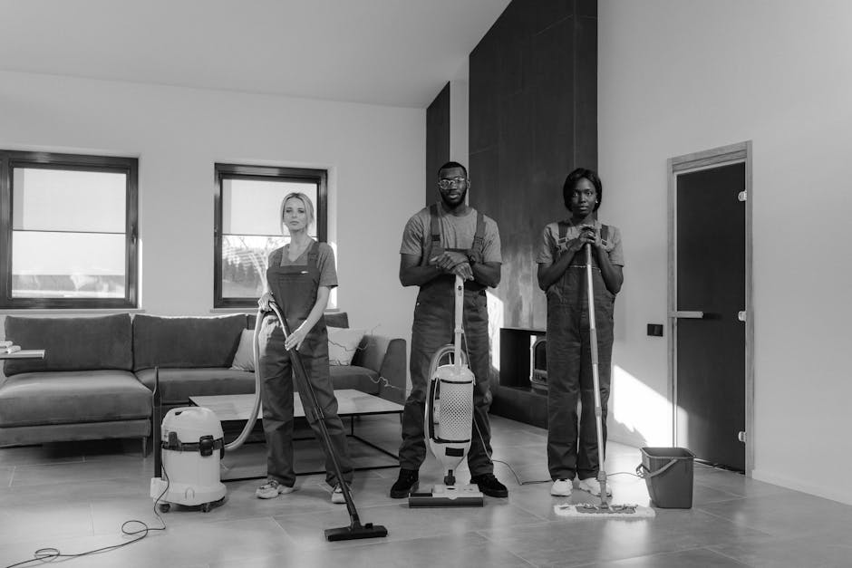 A professional cleaning team consisting of three individuals is performing surface cleaning and deep cleaning in a modern living room. The team members, dressed in work uniforms, are equipped with cleaning tools including vacuum cleaners, mops, and spray bottles. One person is operating a vacuum cleaner on the tiled floor, which appears thoroughly cleaned and shiny, reflecting the natural light coming through two large windows. The other two staff members are using mops and cloths to clean the wooden and tiled surfaces around the room, which are free of dust and grime. The room features a comfortable sofa with cushions, a coffee table, and a sleek fireplace with a dark wooden surround, all well-maintained and free of clutter. Bright natural lighting emphasizes the cleanliness and hygiene of the space. This image showcases the professional domestic cleaning services provided by Cleaners Surrey, aligning with the focus of surface cleaning, sanitisation, and maintenance for same-day house cleaning availability in Woking GU21.