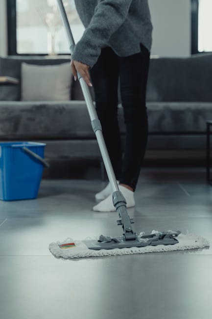 A female cleaner from Cleaners Surrey performing surface cleaning in a modern kitchen. She is dressed in an orange uniform and is using a spray bottle and cloth to wipe down a light-colored, polished wooden dining table. The kitchen features white marble countertops and backsplash, with stainless steel appliances integrated into the cabinetry. Natural light streams through a large window, casting shadows on the wall and creating a bright, calm atmosphere. The scene emphasizes domestic cleaning through thorough sanitisation and surface maintenance of the dining and kitchen surfaces, demonstrating hygienic cleaning processes in a residential setting. The space appears well-maintained and free of dust or stains, reflecting effective deep cleaning by professionals like those at Cleaners Surrey.